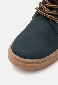 Tmavě zelené semišové tenisky s hnědou gumovou podešví, kontrastními žlutými a námořnickými tkaničkami a texturovaným povrchem na špičce.