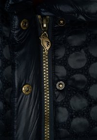 Granatowa pikowana kurtka puchowa z błyszczącym wykończeniem, zdobiona okrągłymi wzorami, złotym zamkiem błyskawicznym i zatrzaskami. Akcent z fakturowanej tkaniny.