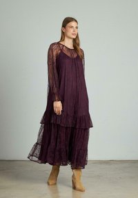 Lungo vestito viola con ruches a strati, realizzato in pizzo. Presenta maniche lunghe e trasparenti e una vestibilità ampia, abbinato a stivali alla caviglia beige.