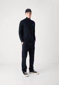 Navy zip-neck sweater with knit texture, paired with matching navy pants. Model wears black cap and black sneakers with white soles.