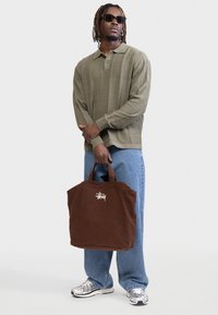 Άνδρας με γυαλιά ηλίου, πράσινο μακρυμάνικο πουκάμισο με υφή, μπλε τζιν και ασημένια αθλητικά παπούτσια, κρατώντας μια μεγάλη καφέ τσάντα ώμου με λευκό λογότυπο.