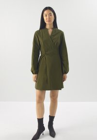 Robe portefeuille vert olive avec de longues manches bouffantes, une ceinture à nouer et une longueur au genou. Portée avec des bottines noires. Texture de tissu lisse.