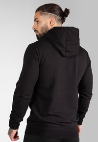 Haut à capuche noir en tissu doux, avec une capuche classique, des manches longues et une coupe ajustée. Vue arrière avec une texture unie.
