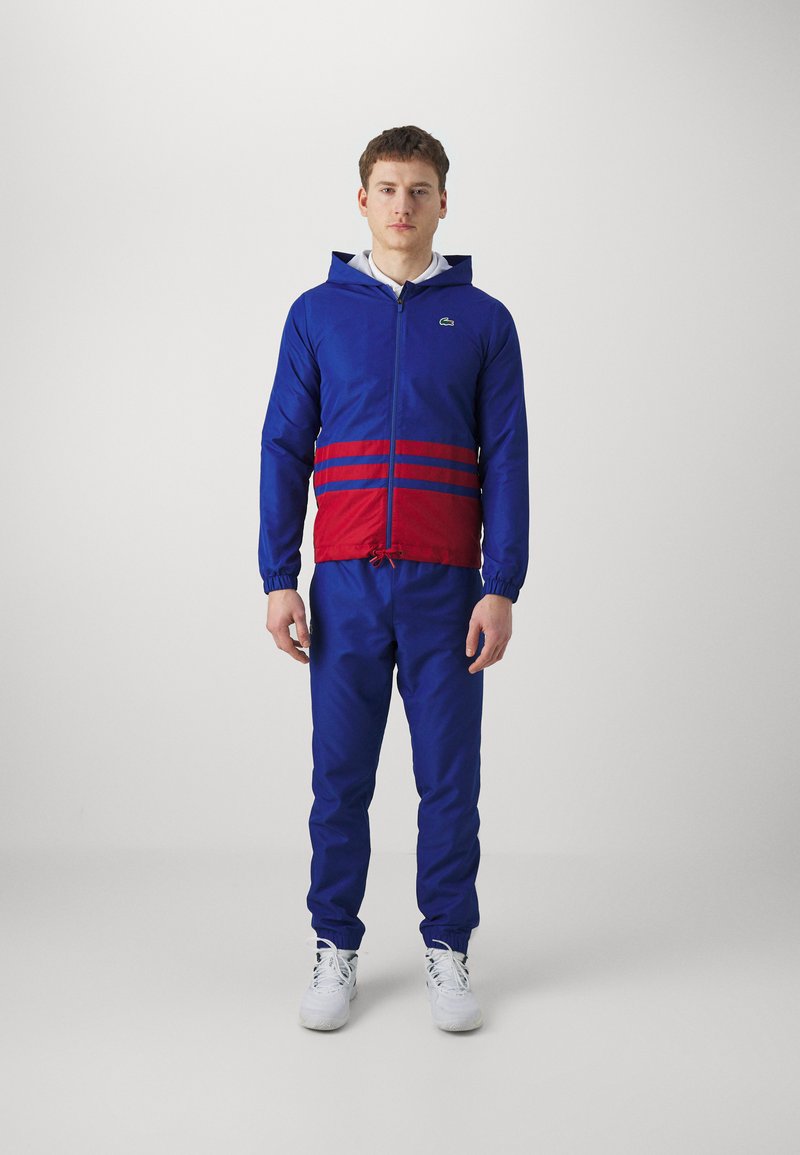 Giacca a zip blu e rossa con cappuccio, caratterizzata da tre strisce rosse in vita. Abbinata a pantaloni blu coordinati e sneaker bianche.