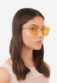 Auksinės metalinės rėmelio akiniai nuo saulės su geltonai tonuotais lęšiais, turintys geometrinę formą ir minimalų detalių kiekį. Modelis turi ilgus, tiesius rudus plaukus.