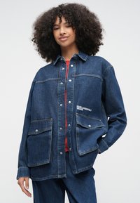 Kobieta o kręconych włosach, ubrana w za dużą ciemnoniebieską kurtkę jeansową i dopasowane dżinsy, stoi z jedną ręką w kieszeni i lekkim uśmiechem.