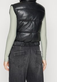 Osoba w bezrękawniku z czarnej skórzanej puchówki, nosząca długi jasnobrązowy top i ciemnoszare dżinsy, pokazana z tyłu.