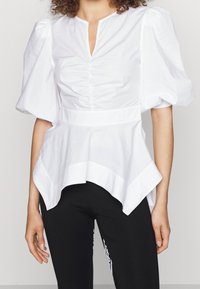 Blusa bianca con maniche corte a sbuffo, scollatura a V e dettagli arricciati sul davanti; presenta un orlo asimmetrico e tessuto in cotone morbido.