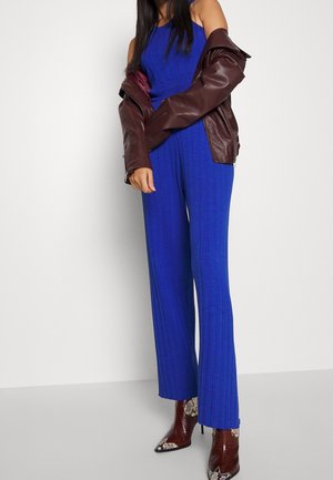 Mujer con un mono sin mangas azul acanalado, chaqueta de cuero burdeos caída de los hombros y botines marrones con punta y estampado de piel de serpiente.