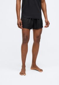 Mustad mustad ujumispüksid tekstuurilise mustriga, paelast vöökoht ja külje lõhikud, koos musta T-särgiga, seljas inimesel, kes seisab paljajalu.