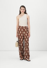 Beige topp med én skulder; brune vide bukser med kremfargede bladmønstre; holder en teksturert kremfarget veske; svarte sandaler; glatt stoff.