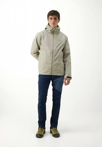 Veste imperméable beige avec capuche et fermeture éclair, associée à un pantalon bleu marine et des chaussures de randonnée vertes. La veste comporte un logo sur la poitrine.