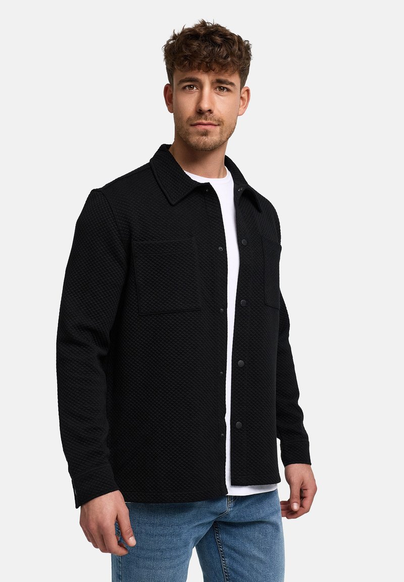 Junger Mann trägt eine schwarze, strukturierte Knopfleistenjacke über einem weißen Hemd und blauer Jeans, steht vor einem schlichten hellen Hintergrund.