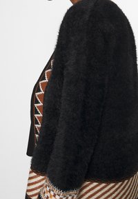 Persona vistiendo un suéter negro de lana sobre una blusa y falda de patrones marrón, blanco y negro con diseños geométricos, mostrado de lado.