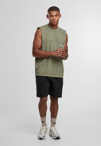 Uomo atletico in piedi con le mani incrociate, indossa una maglietta senza maniche color oliva, pantaloni corti neri, calzini beige e scarpe da corsa argentate su uno sfondo semplice.