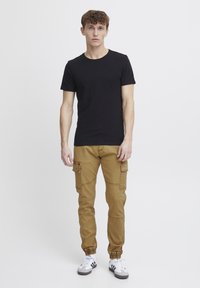 Czarny t-shirt z krótkim rękawem, beżowe spodnie cargo z bocznymi kieszeniami, elastyczne mankiety i białe sneakersy w czarne paski. Jednolity tło.