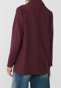 Burgundy bleiseri, jossa on kauluksen muotoinen kaulus, pitkät hihat ja nappikoristeet. Bleiserissä on sileä pinta ja muotoiltu istuvuus.