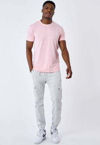Uomo in piedi con le mani rilassate, indossa una maglietta rosa chiaro, pantaloni cargo grigio chiaro e sneakers bianche con dettagli neri.