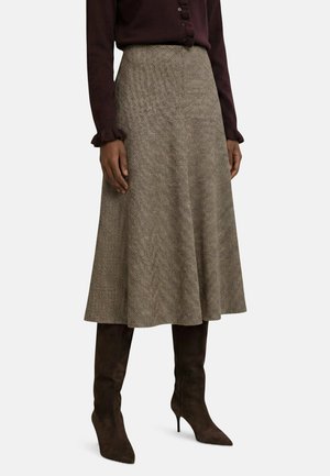 Femme portant une jupe midi texturée taille haute associée à des bottines à talons brun foncé et un chemisier à manches longues avec des détails de boutons.