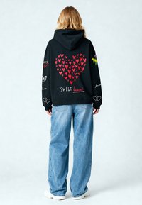 Sudadera negra con un gran gráfico de corazón rojo, texto que dice "SWEET heart" y diseños coloridos en las mangas. Combinada con unos vaqueros de color azul claro.