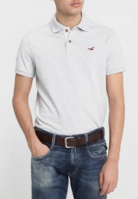 Lys grå polo-shirt med to brune knapper ved kraven og et lille rødt logo på brystet, parret med blå jeans og et brunt bælte.