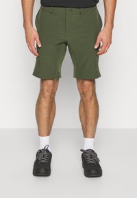 Pantalones cortos verde oliva hechos de una tela ligera, con una cintura estándar, bolsillos laterales y un dobladillo plegable. Combinados con zapatillas negras y calcetines blancos.