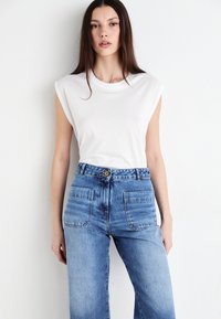 Camiseta sin mangas blanca combinada con unos vaqueros de mezclilla azul claro de cintura alta, que cuentan con un cierre de botón y dos bolsillos traseros.