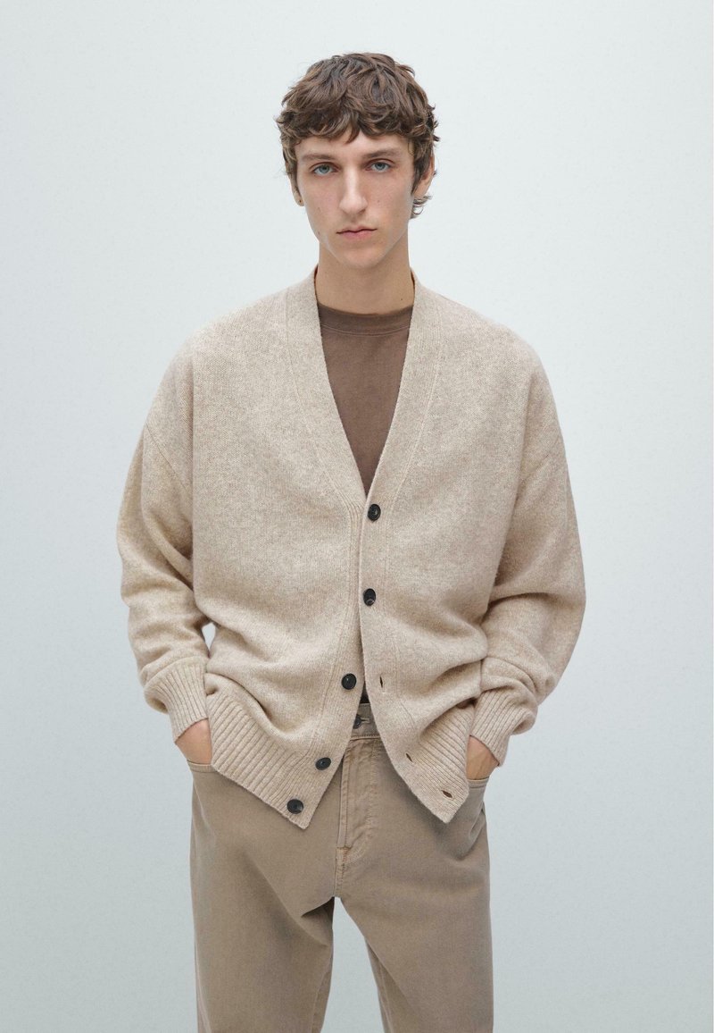 Beżowy sweter na drutach z dekoltem w serek, ściągaczowymi mankietami i brązowymi guzikami. Nosi się go na brązową koszulkę, w zestawie z beżowymi spodniami. Jednolite tło.