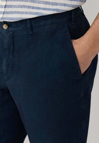 Primer plano de moda masculina que muestra pantalones azul oscuro con una mano en el bolsillo lateral y parte de una camisa a rayas metida dentro.