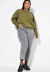 Sudadera verde oliva con cuello acanalado, texto "VIBRACIONES DE FIN DE SEMANA", combinada con jeans ajustados gris claro y mocasines negros gruesos.