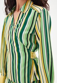 Blouse portefeuille rayée verte et jaune avec col, manches longues, détail de nouage sur le côté, et texture lisse. Lignes verticales de largeurs variées.