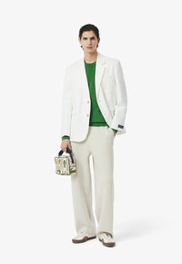 Weißer Blazer, grüner Rundhalspullover, beiger, weiter Hosenanzug, hält eine gemusterte Tasche mit Stickerei und schwarzen Akzenten, sowie cremefarbene Schuhe.