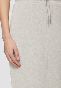 Helles graues Sweatshirtmaterial mit strukturierter Oberfläche, ausgestattet mit einem Kordelzugverschluss und Seitentaschen, teilweise von einer Hand verdeckt.