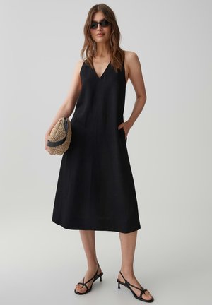 Frau trägt ein schwarzes ärmelloses Midikleid, schwarze Riemchenabsätze, Sonnenbrille, hält eine beige gewebte Clutch, steht vor einem schlichten hellen Hintergrund.