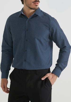 Homme portant une chemise bleu marine à boutons avec un petit motif à carreaux blancs et un pantalon noir, une main dans la poche, tourné légèrement vers la droite.