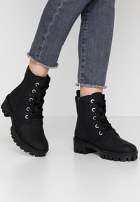 Stivaletti neri alla caviglia con finitura testurizzata, parte anteriore con lacci, occhielli metallici, zipper laterale e suola in gomma spessa. Jeans con orli sfilacciati.