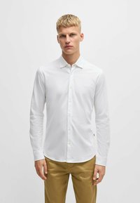 Chemise blanche à manches longues avec un bouton, faite d'un tissu lisse, présentant un col classique et un petit logo sur la poitrine. Associée à un pantalon beige.