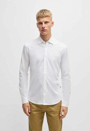 Chemise blanche à manches longues avec un bouton, faite d'un tissu lisse, présentant un col classique et un petit logo sur la poitrine. Associée à un pantalon beige.