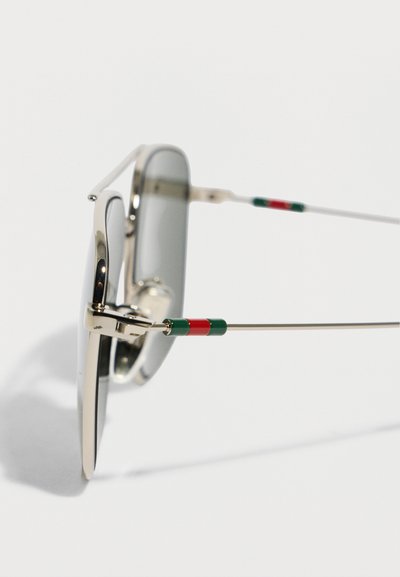 Κοντινό πλάνο ορθογώνιων γυαλιών ηλίου με ασημένια στεφάνη και λεπτούς βραχίονες που φέρουν κόκκινες και πράσινες ρίγες, σε λευκό φόντο.