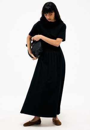 Žena s dlhými čiernymi vlasmi, oblečená v čiernom tričku s krátkym rukávom a dlhej sukni, drží čiernu tkanú kabelku, na nohách má hnedé baleríny, na bielom pozadí.