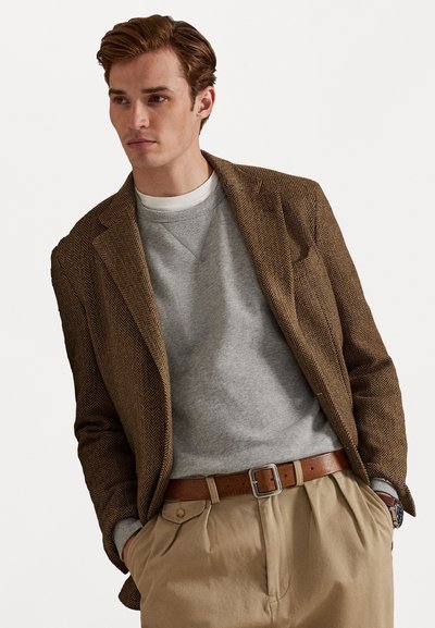 Blazer castanho de espinha de peixe sobre um sweatshirt cinza, calças pregueadas em tonalidade bege e um cinto de couro castanho com uma fivela quadrada prateada.