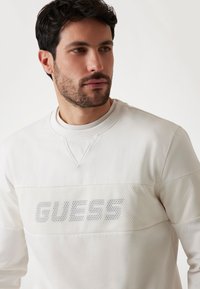 Bijela majica s dugim rukavima koja sadrži mrežastu površinu s natpisom "GUESS". Okrugli izrez, glatka tekstura i sportski dizajn.