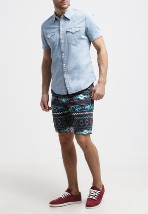 Man in een lichtblauwe spijkerhemd met korte mouwen, met gepatterneerde kniebange shorts in turkoois, zwart, rood, en rode sneakers met witte veters.