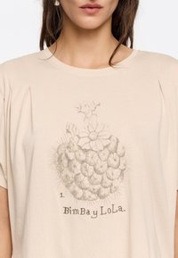 Top beige a maniche corte caratterizzato da una grafica di un cactus con fiori e la scritta "BimBAY LoLa." Realizzato in materiale morbido e leggero.