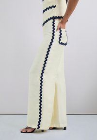 Flared trousers in a light yellow fabric featuring navy zigzag trim along the sides, with a side pocket and a relaxed fit.