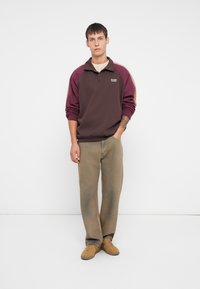 Rjav in burgundski polo pulover s ovratnikom, v kombinaciji s svetlobruna hlačami in bež športnimi copati, na modelu, ki stoji pred belo ozadjem.