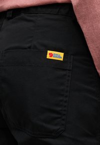 Poche arrière d'un pantalon noir avec une étiquette logo Fjällräven jaune, associée à un tissu rose clair visible au-dessus de la ceinture.
