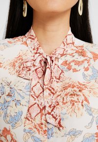 Blommig blus med en ljus bakgrund och mönster i röd, blå och persika, avslutad med en rosett vid kragen. Slät tygtextur.