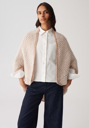 Châle tricoté rose pâle avec des côtés ouverts, motif texturé, porté sur une chemise blanche à boutons, associé à un pantalon large bleu marine.