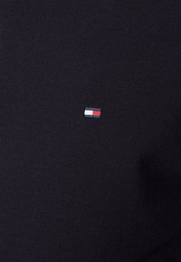 Marinblå polo skjorta med en liten broderad logotyp och röda, vita och blå rektangulära accenter på bröstet. Texturerat stickat tyg.